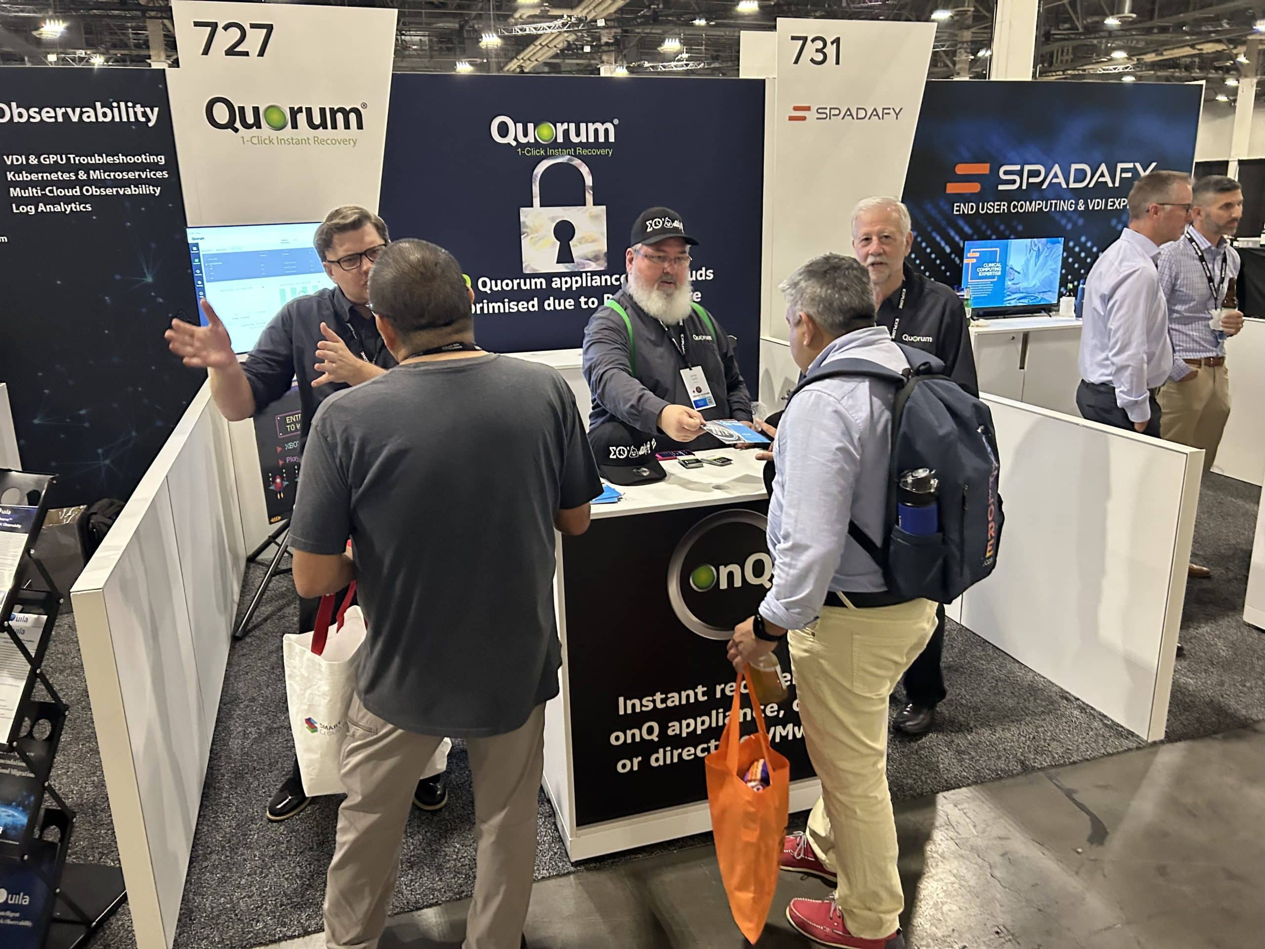 A group of people are interacting at a tradeshow booth for Quorum, a company providing IT disaster recovery solutions. The booth has monitors and promotional materials. Nearby, there are other booths labeled "727" and "731" showcasing different tech companies.