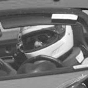 A person wearing a helmet is seated in a car, visible through the open window. The helmet is white with a clear visor, and the individual appears to be looking forward. The image is in black and white, capturing a focused moment.
