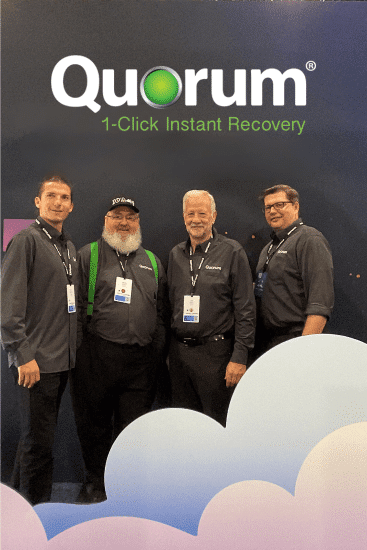 Four men are standing and smiling under a sign that reads "Quorum 1-Click Instant Recovery." They are all wearing matching dark grey shirts with the Quorum logo and conference badges. One individual is wearing a green backpack and a black "IOT Village" hat.