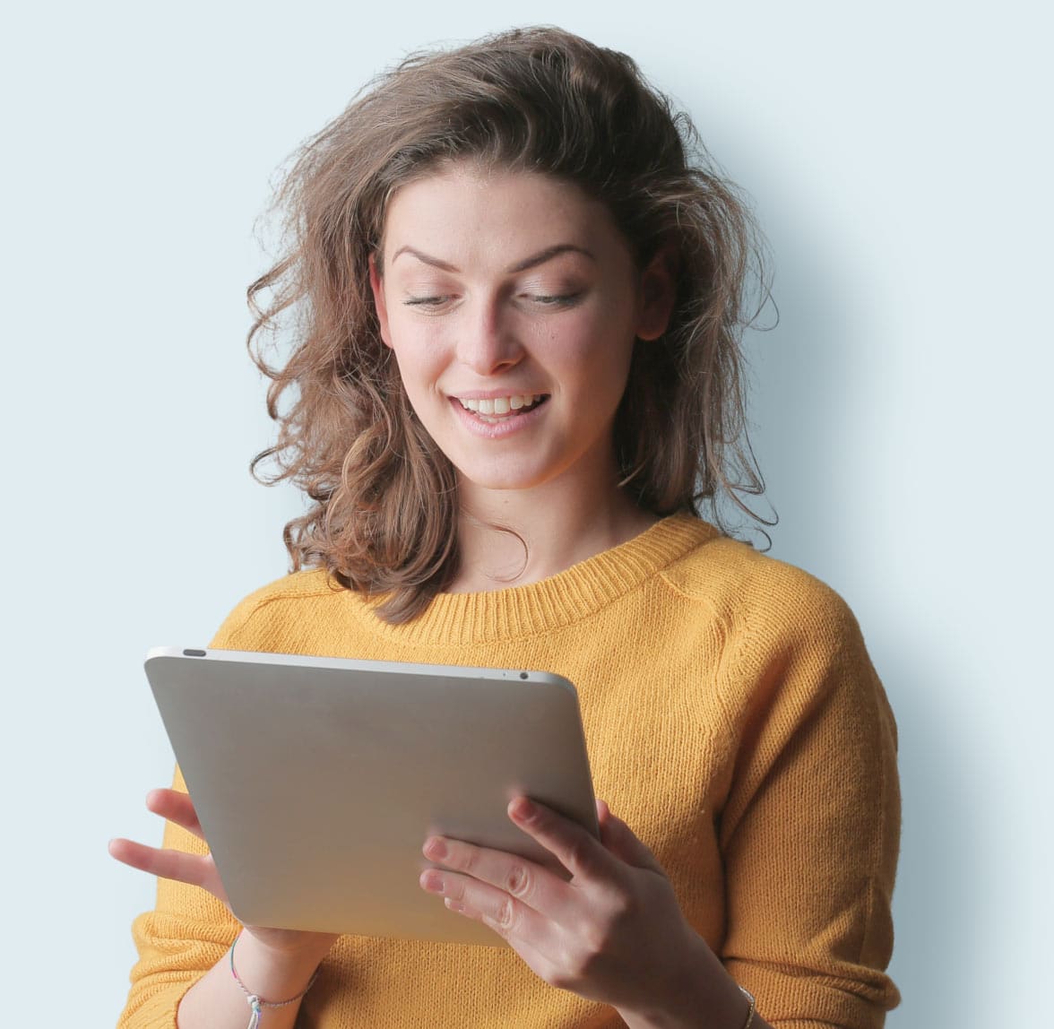 A person with wavy brown hair is smiling while looking at a tablet. They are wearing a yellow sweater and standing against a light blue background.